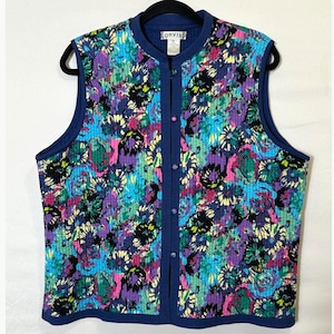 May include: A sleeveless quilted vest by Orvis featuring a vibrant floral pattern in shades of blue, purple, green, yellow, and black. The vest has a solid blue ribbed collar and trim, a front button closure, and a label reading Orvis.