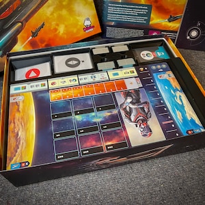 May include: An open box game with a space theme. The box contains a game board with a planet, stars, and a spaceship. There are also cards, dice, and other game pieces.