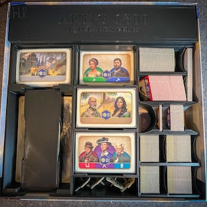 Könnte beinhalten: Eine schwarze Spielbox mit dem Text "ANNO 1800 POPULATION CUBES" auf dem Deckel. Die Box ist in Fächer unterteilt, die Spielkarten und illustrierte Karten mit Personen und Stadtlandschaften enthalten. Die Spielkarten sind in Stapeln.