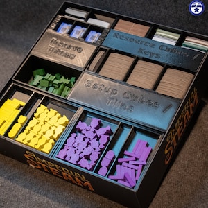 Puede incluir: Un organizador negro con compartimentos para guardar las piezas y fichas del juego. El organizador está etiquetado como "Imperial Steam" y tiene compartimentos etiquetados como "Reserve Tokens", "Resource Cubes/Keys", "Setup Cubes/Tiles". Los compartimentos contienen varias piezas de juego de colores diferentes, incluyendo amarillo, verde, morado y azul.