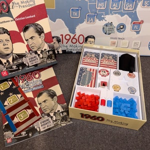 May include: A board game titled "1960: The Making of the President" with a game board, cards, and playing pieces. The game board is a map of the United States with states labeled. The cards are red, white, and blue with the text "1960" on them. The playing pieces are red and blue cubes.