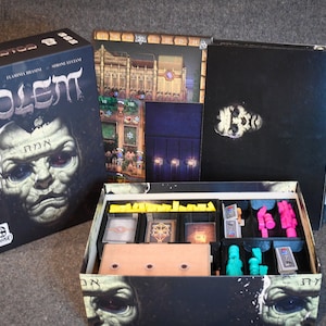 Puede incluir: Se muestra el juego de mesa "Golem", con una caja con un diseño oscuro y detallado de un rostro. Dentro de la caja hay tableros, cartas y piezas de juego, lo que sugiere un juego de estrategia para jugar en la mesa.