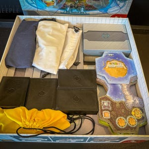 May include: An open game box containing various components, including a blue and white game board, four black storage boxes, three white drawstring bags, and a yellow drawstring bag. The game board features a blue and white island with a yellow building and the text "The Isle of Cats".