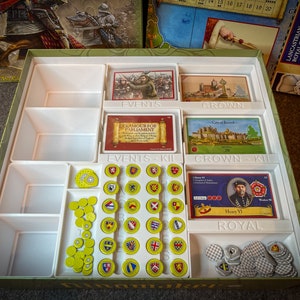 Può includere: Un organizzatore in plastica bianca con scomparti per riporre pezzi di gioco e carte. L'organizzatore è etichettato con "Eventi", "Corona", "Kit Eventi", "Kit Corona" e "Reale". Ci sono anche diversi pile di gettoni rotondi nell'organizzatore.