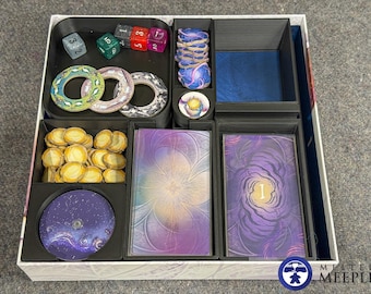 May include: A black plastic board game organiser insert containing various game components. It features compartments holding multi-coloured dice, circular tokens, stacks of yellow coins, a purple circular disc, and two decks of cards with purple floral designs.