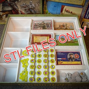 Può includere: Un inserto in plastica bianca per un gioco da tavolo con scomparti per gettoni, carte e altri pezzi di gioco. L'inserto è etichettato "Eventi", "Corona", "Kit Corona" e "Reale".