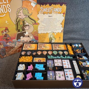 Könnte beinhalten: Das Brettspiel "Sweet Lands" mit einer farbenfrohen Box, die eine Illustration eines Charakters mit einem Dessert zeigt. Das Spiel enthält Spielkarten, Spielsteine und verschiedene Spielteile.