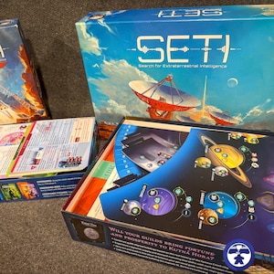 May include: Several board game boxes are displayed, featuring the title "SETI" and imagery of space and rockets. One box shows a rocket launch, while another depicts a satellite dish. The game boards inside are visible.