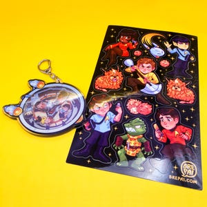 May include: A keychain and sticker sheet featuring cartoon illustrations of characters and creatures. The keychain is a clear, circular design with a spaceship outline. The sticker sheet has various character stickers on a black background.