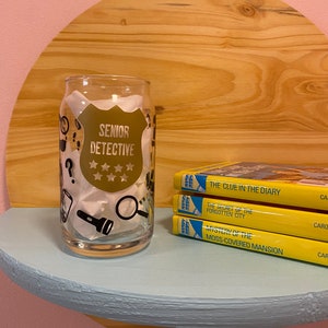 May include: A glass cup with a design of a detective badge that says "Senior Detective" with five stars. The cup is sitting on a light blue shelf with three yellow paperback books stacked on top of it. The books are from the Nancy Drew series and the titles are "The Clue in the Diary", "The Secret of the Forgotten City", and "Mystery of the Moss-Covered Mansion".