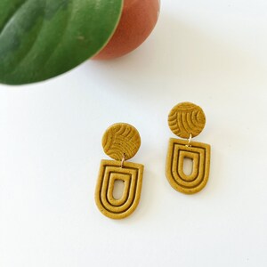 May include: A pair of mustard yellow dangle earrings with a geometric design. The earrings feature a small round disc with a textured pattern and a larger, curved, open-ended shape with three concentric rings.