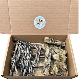 May include: A cardboard box containing a selection of dog treats. The box is open, revealing twisted, braided, and flat dried chews in grey and tan hues. A circular sticker on the lid displays the text "Dried Goodies" and "100% Natural Dog Treats."