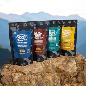 Op de afbeelding: Vier zakjes met bottenbouillon voor honden, gelabeld met verschillende smaken: eland, rund, eend en kip. De zakjes zijn zwart met poot afdrukken en dragen de merknaam "Nova Dog Chews".