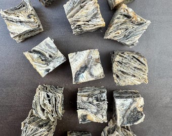 May include: A close-up of a pile of dried fish treats for dogs. The treats are square-shaped and have a light brown and dark gray color. The treats are arranged on a brown surface.