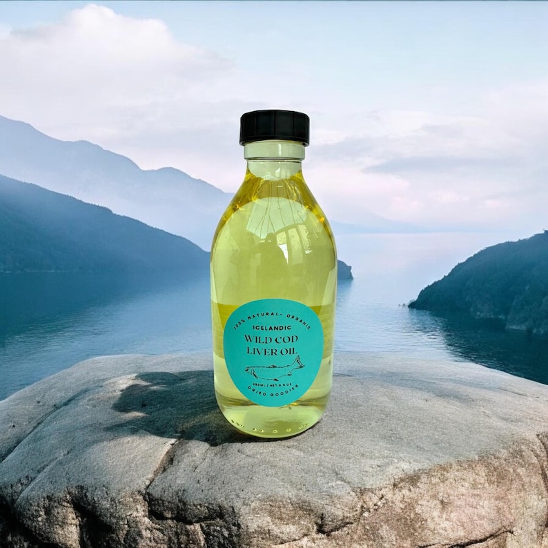 Puede incluir: Una botella de vidrio transparente de aceite de h&iacute;gado de bacalao salvaje island&eacute;s, con una tapa negra, sentada sobre una roca con vista a un cuerpo de agua y monta&ntilde;as en el fondo.