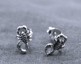 May include: A pair of silver scorpion stud earrings. Each earring has a detailed scorpion design with a textured body and curved tail. The earrings are shown on a grey fabric surface.