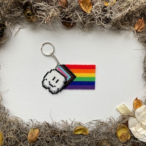 May include: A rainbow pride flag keychain with a white pixelated smiley face charm. The keychain is made of colorful plastic beads.