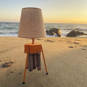 May include: A table lamp with a light-colored, textured fabric lampshade and a hexagonal wooden base. The base is supported by three thin, angled wooden legs and features a small metal switch on the front, with several vertical spools of thread.