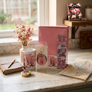 May include: A pink gift box set containing a white ceramic mug, a circular keychain, a square sticker, and a tall bookmark, all featuring the text I AM NOT MOST GIRLS I AM A VETERAN and a graphic of combat boots. A leather journal and compass are nearby.