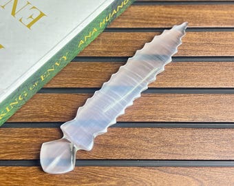 Könnte beinhalten: Ein durchscheinender, hellfarbiger Kristalldolch mit gezackter Kante, der auf einer Holzoberfläche neben einem Buch mit dem Titel "King of Envy" von Ana Huang liegt. Der Dolch hat einen rechteckigen Griff.