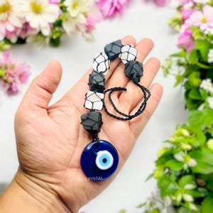 Może przedstawiać: Czarne i białe koraliki kryształowe nawleczone na czarny sznurek z niebieskim amuletem w kształcie oka na końcu. Amulet ma białe oko i białą źrenicę. Koraliki mają geometryczny kształt i są owinięte czarną siatką. Amulet wisi na dłoni.