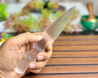 Könnte beinhalten: Ein klarer, durchscheinender Kristalldolch mit spitzer Klinge und dekorativem Griff. Der Dolch wird in einer Hand gehalten, wodurch seine glatte, polierte Oberfläche hervorgehoben wird. Der Hintergrund ist unscharf.