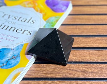 Könnte beinhalten: Ein glänzender, schwarzer Kristall in Pyramidenform liegt auf einem Buch mit dem Titel "Crystals for Beginners". Das Buch hat einen gelben Einband mit bunten Kristallen. Die Pyramide befindet sich auf einer Holzoberfläche.