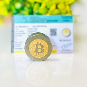 Può includere: Un oggetto rotondo grigio e oro a tema Bitcoin con il simbolo Bitcoin al centro. L'oggetto è su una superficie bianca con uno sfondo sfocato. Il simbolo Bitcoin è dorato e l'oggetto ha un bordo grigio.
