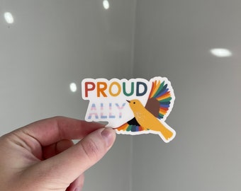 May include: A sticker with the words "PROUD ALLY" in colorful letters. The sticker features a yellow bird with rainbow-colored wings. The bird is in flight, with its wings spread wide. The background is a light gray.