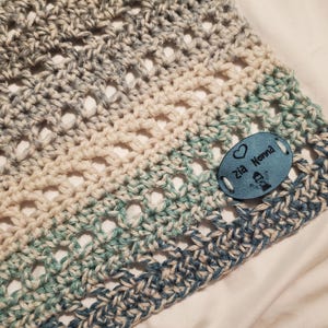 May include: A crocheted baby blanket in shades of blue, white, and green with a blue oval tag that says "Zia Nonna" with a heart and a small illustration of a person.