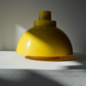 Könnte beinhalten: Eine gelbe, kuppelförmige Lampe aus Kunststoff mit einem kleineren, gelben Kunststoffzylinder oben. Die Lampe steht auf einer weißen Oberfläche.