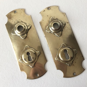 May include: Two antique brass door plates with keyhole and knob openings. Each plate has a rectangular shape with decorative edges and is secured with small screws. The brass has a slightly aged patina.