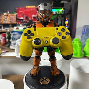 May include: A figurine of a futuristic soldier holding a yellow PlayStation 4 controller. The soldier is orange and gray, standing on a black base with a gold Xbox logo. The controller has black buttons and a PlayStation logo.