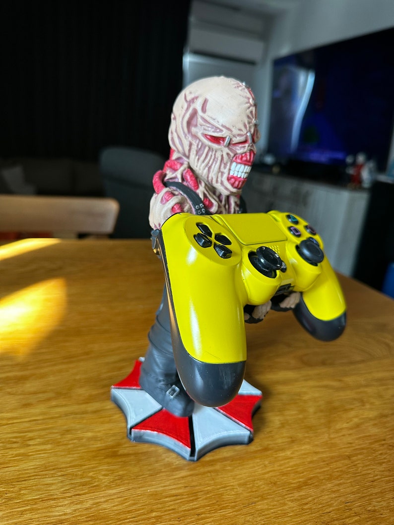 Pode incluir: Um controlador de videogame amarelo e preto segurado por uma figura de uma criatura monstruosa com um logotipo de guarda-chuva vermelho e branco na base.