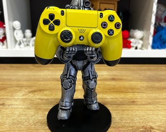 Puede incluir: Una figura de robot plateado sosteniendo un mando de PlayStation 4 amarillo. El robot está de pie sobre una base circular negra.