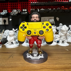 May include: A yellow PlayStation 4 controller is held by a figurine of a soccer player wearing a red and white uniform with the number 11 on the front. The figurine is standing on a black base with a white logo that says "EST. 1892".