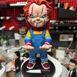 Puede incluir: Figura de muñeco Chucky con cabello naranja, ojos azules y detalles faciales cosidos. El muñeco lleva un peto azul sobre una camisa a rayas y zapatillas rojas. Se encuentra sobre una base negra con un cuchillo.