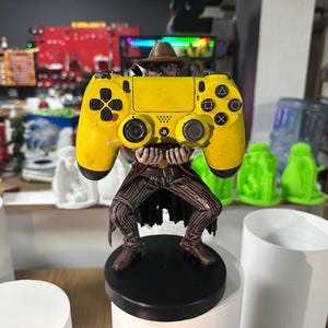 May include: A yellow video game controller statue held by a figure in a cowboy hat and vest. The statue is on a black circular base. The controller has black buttons and a directional pad.