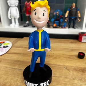 May include: A figurine of the Fallout character Vault Boy, standing on a black base. The figure is wearing a blue jumpsuit with yellow accents and has blonde hair. The base reads "VAULT-TEC" in white letters.