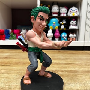 May include: A figurine of a character with green hair, wearing a white tank top, gray pants, and brown sandals. The figure is posed with arms outstretched, holding three swords. The figurine is on a black circular base.