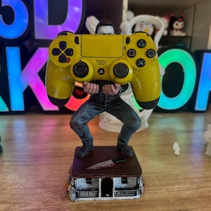 May include: A yellow PlayStation 4 controller held by a figurine of a person in jeans and a black shirt. The figurine stands on a small house with a bloody knife. The background has neon letters.