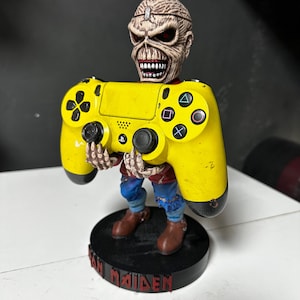 Puede incluir: Una figura de Eddie, la mascota de la banda de heavy metal Iron Maiden, sostiene un mando de PlayStation 4 amarillo. La figura lleva un atuendo rojo y azul y está de pie sobre una base negra con el nombre de la banda escrito en rojo.