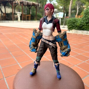 May include: A collectible action figure of a female character with pink hair, wearing a white top, brown pants, and blue and gold gauntlets. The figure is standing on a brown surface.