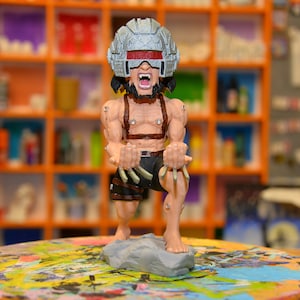 May include: A hand-painted figurine of a superhero character with a silver helmet, red visor, and a snarling expression. The figurine is posed in a dynamic stance with claws extended, and is standing on a gray rock base. The figurine is on a colorful paint palette.