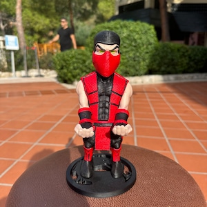 Könnte beinhalten: Eine rote und schwarze Figur eines Ninja-Charakters mit einer roten Maske und grünen Augen. Die Figur ist in einer hockenden Position mit den Händen in Fäusten dargestellt. Die Figur steht auf einem schwarzen Sockel mit einem kreisförmigen Design.