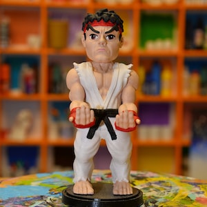 以下が含まれることがあります： 白い道着に黒いベルトをつけた格闘技のキャラクターのフィギュア。キャラクターは赤いヘッドバンドを付けており、両手を上げて戦いの姿勢をとっている。