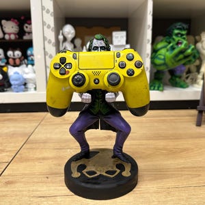May include: A figurine of the Joker holding a yellow video game controller. The Joker is wearing a purple suit and green vest. The base is black with a gold design. Other figurines are visible in the background.