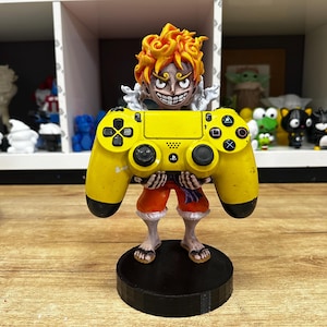 May include: A yellow and black video game controller stand featuring a cartoon character holding the controller. The character has spiky orange hair and a yellow and black outfit.