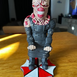 Pode incluir: Uma figura preta e vermelha de um monstro com um rosto costurado e um traje preto. A figura est&aacute; em p&eacute; sobre uma base preta e branca com o logotipo de um guarda-chuva.