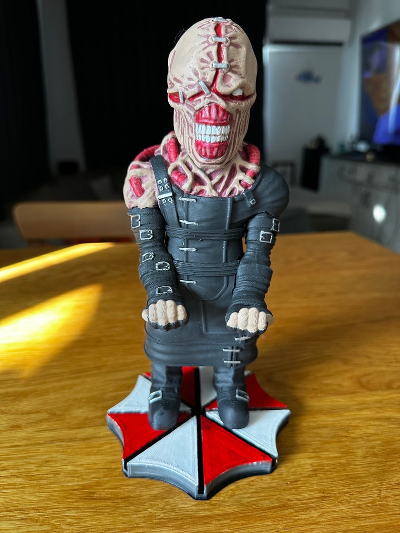 Pode incluir: Uma figura preta e vermelha de um monstro com um rosto costurado e um traje preto. A figura est&aacute; em p&eacute; sobre uma base preta e branca com o logotipo de um guarda-chuva.